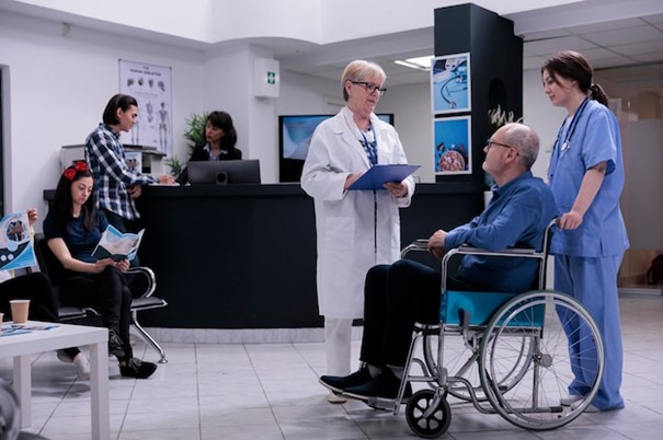 Free photo retired person using wheelchair talking with senior doctor for appointment while being helped by professional nurse in private clinic. older man living with disability in busy hospital reception.